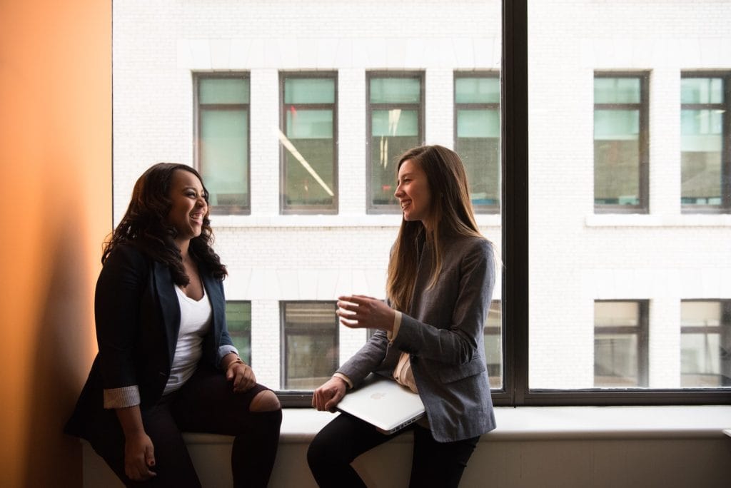 Two women happily chatting while sitting on the window overlooking another building.