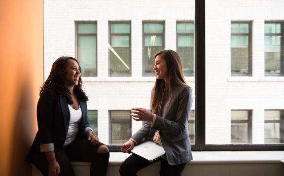 Two women happily chatting while sitting on the window overlooking another building.