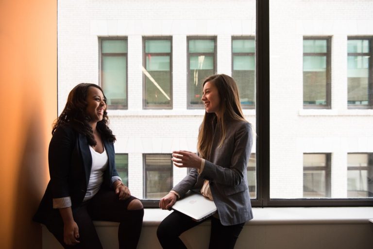 Two women happily chatting while sitting on the window overlooking another building.